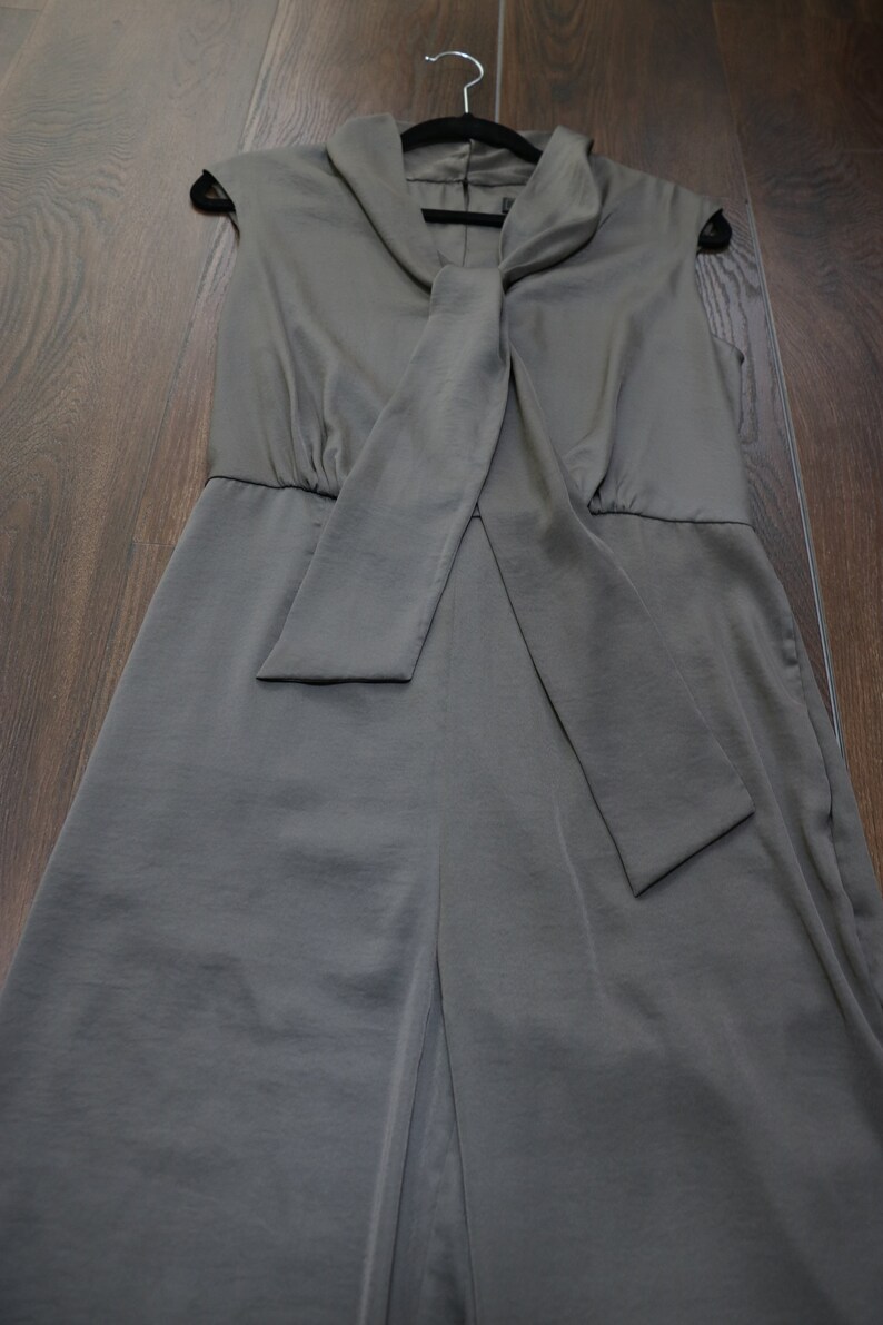 Puede incluir: Mono sin mangas verde oliva con cuello drapeado y detalle de lazo a juego. La prenda cuelga de una percha negra. La tela tiene un acabado liso, satinado. El fondo es un suelo de madera.