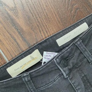 May include: Close-up of dark grey denim jeans. The jeans have a button closure and a small tag that reads "SCRIPT Size 27". A second tag reads "Pilcro and the letterpress". A third tag reads "Beaded Waist Rise".