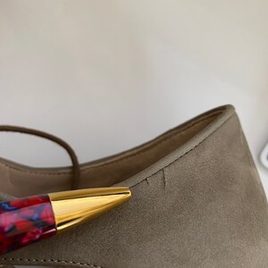May include: Close-up of a taupe suede sandal with a gold-tipped pen. The sandal has a strappy design and the brand name "Valentino Garavani" is visible. The pen has a red and blue patterned barrel.