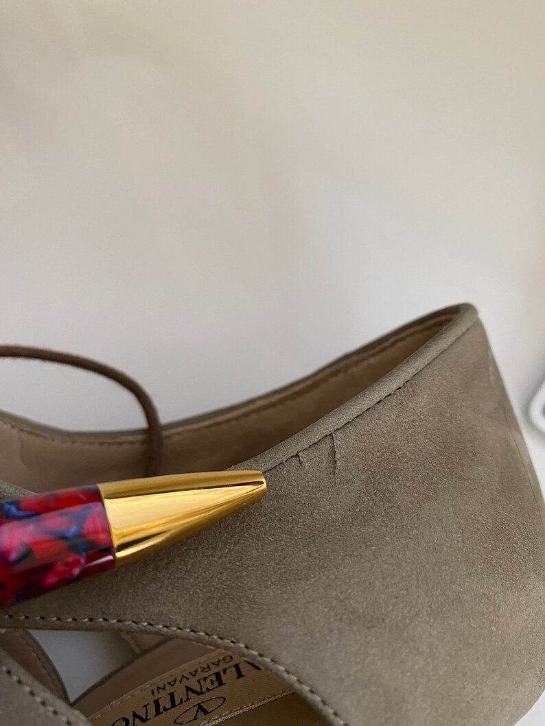 May include: Close-up of a taupe suede sandal with a gold-tipped pen. The sandal has a strappy design and the brand name "Valentino Garavani" is visible. The pen has a red and blue patterned barrel.