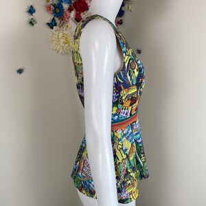 May include: A sleeveless top with a vibrant print of buildings and abstract designs in yellow, blue, and green. The top has a fitted bodice and a flared peplum, with an orange and green band at the waist. The garment is displayed on a mannequin.