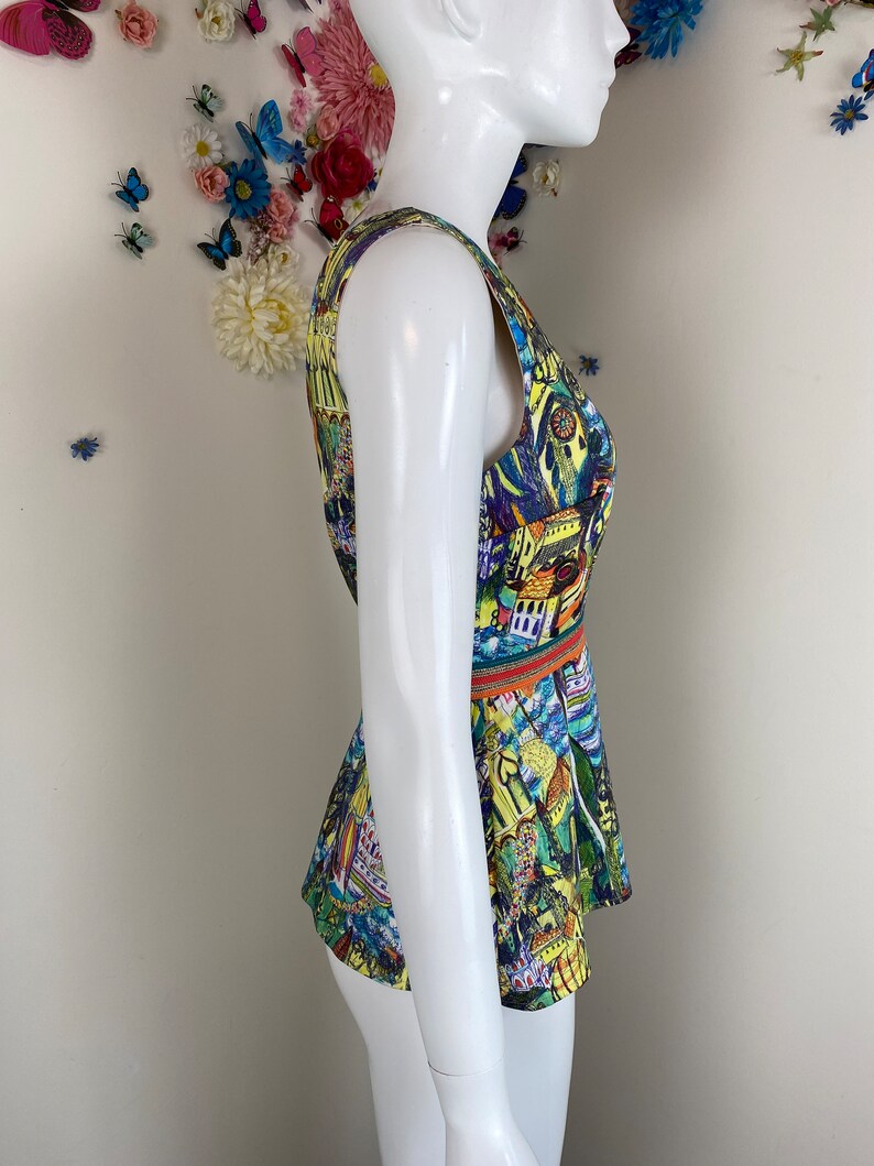 May include: A sleeveless top with a vibrant print of buildings and abstract designs in yellow, blue, and green. The top has a fitted bodice and a flared peplum, with an orange and green band at the waist. The garment is displayed on a mannequin.