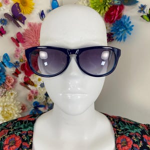 May include: Dark navy blue sunglasses with a rounded rectangular frame and gradient tinted lenses. The sunglasses are displayed on a smooth, featureless white mannequin head, showcasing the deep blue plastic frame and the dark lens color.