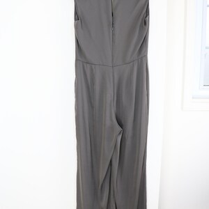 Puede incluir: Mono gris sin mangas con cuello alto, colgado de una percha negra. La parte trasera del mono tiene cierre de cremallera y un bot&oacute;n en el cuello. La tela tiene un brillo sutil.