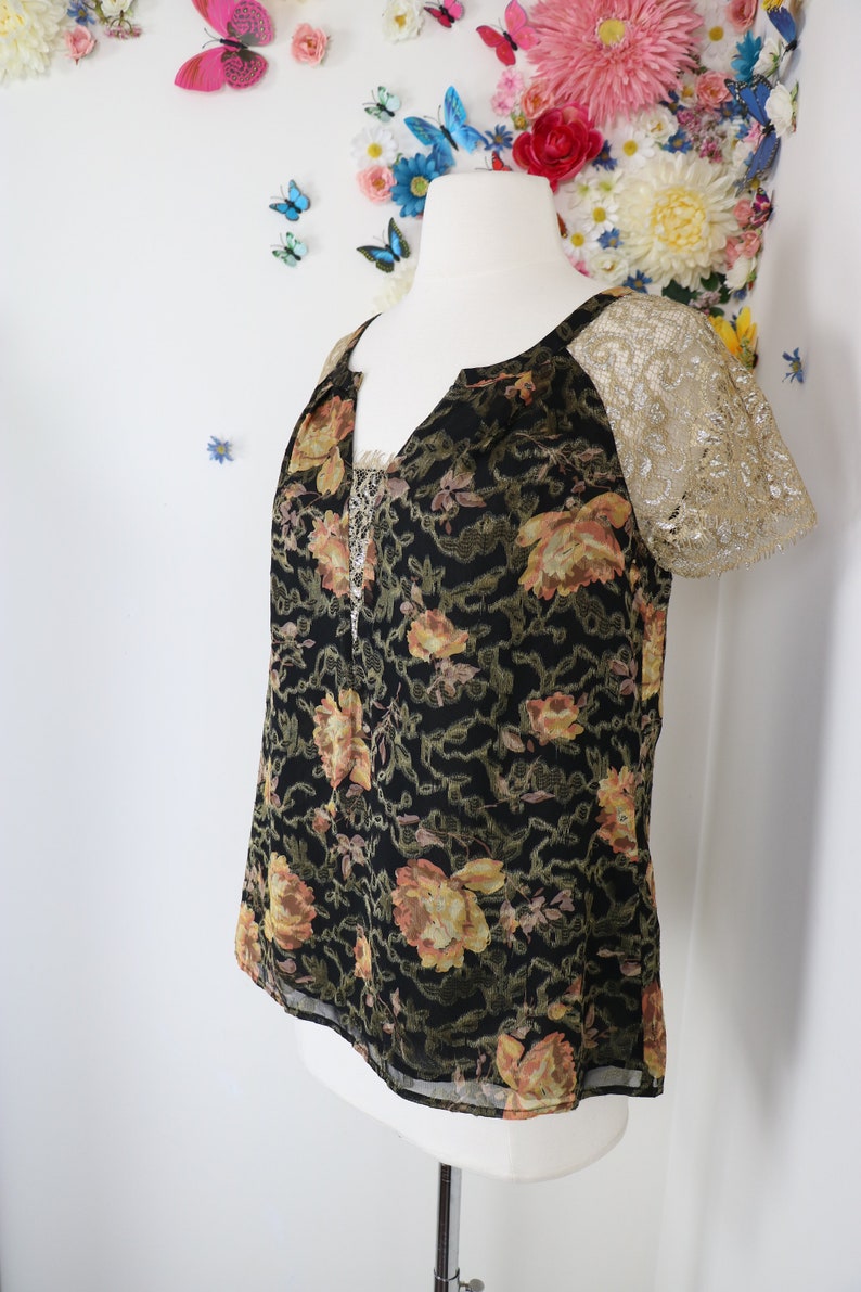 Puede incluir: Top negro con estampado floral y mangas de encaje dorado. El top presenta un escote en V y un delicado ribete de encaje. El estampado floral incluye tonos amarillos y naranjas.