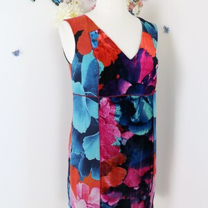 Pode incluir: Vestido sem mangas, na altura do joelho, com decote em V, exibido em um manequim. O vestido apresenta um vibrante padr&atilde;o floral em tons de vermelho, azul, rosa e preto. Uma fina borda vermelha acentua a cintura. O fundo inclui decora&ccedil;&otilde;es florais e borboletas.