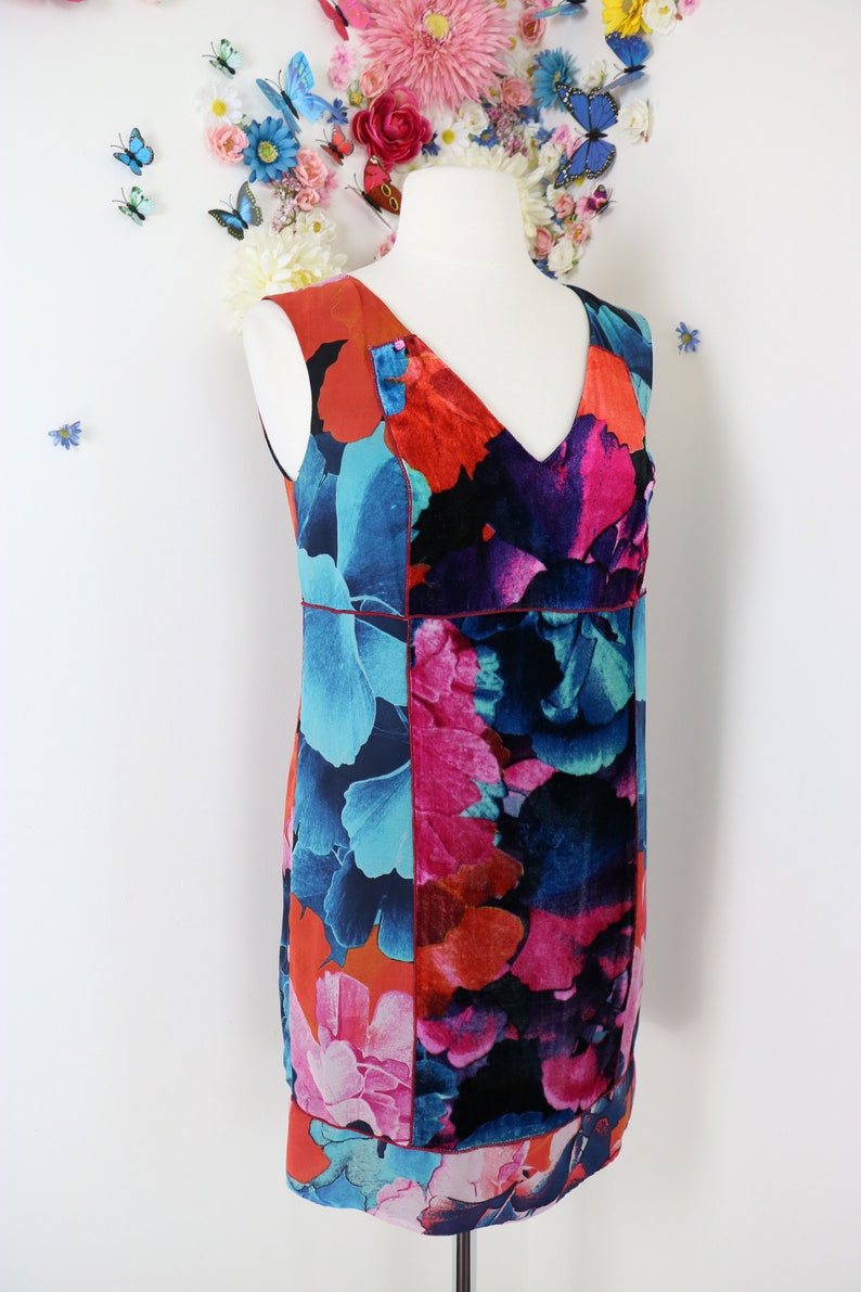 Pode incluir: Vestido sem mangas, na altura do joelho, com decote em V, exibido em um manequim. O vestido apresenta um vibrante padr&atilde;o floral em tons de vermelho, azul, rosa e preto. Uma fina borda vermelha acentua a cintura. O fundo inclui decora&ccedil;&otilde;es florais e borboletas.