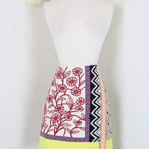 May include: A colorful, patterned skirt displayed on a mannequin. The skirt features a white background with red floral embroidery, a yellow band, and a black and purple checkered hem with pom-pom trim. Various patterned panels add to the design.