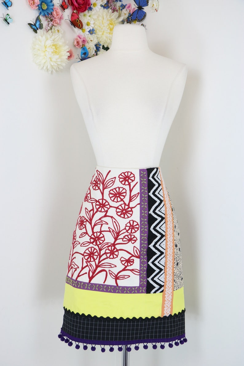 May include: A colorful, patterned skirt displayed on a mannequin. The skirt features a white background with red floral embroidery, a yellow band, and a black and purple checkered hem with pom-pom trim. Various patterned panels add to the design.
