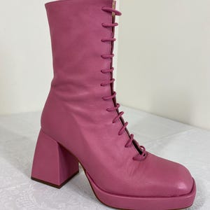 May include: A pink, lace-up boot with a square toe and a chunky, block heel. The boot features a platform sole and a mid-calf height. The laces are the same pink color as the boot. The heel measures approximately 4 inches.