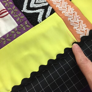 May include: Close-up of a patchwork textile with various textures and colors. Features include black and white beaded chevron patterns, orange and white patterned ribbon, and a bright yellow fabric. A black grid pattern fabric is visible.