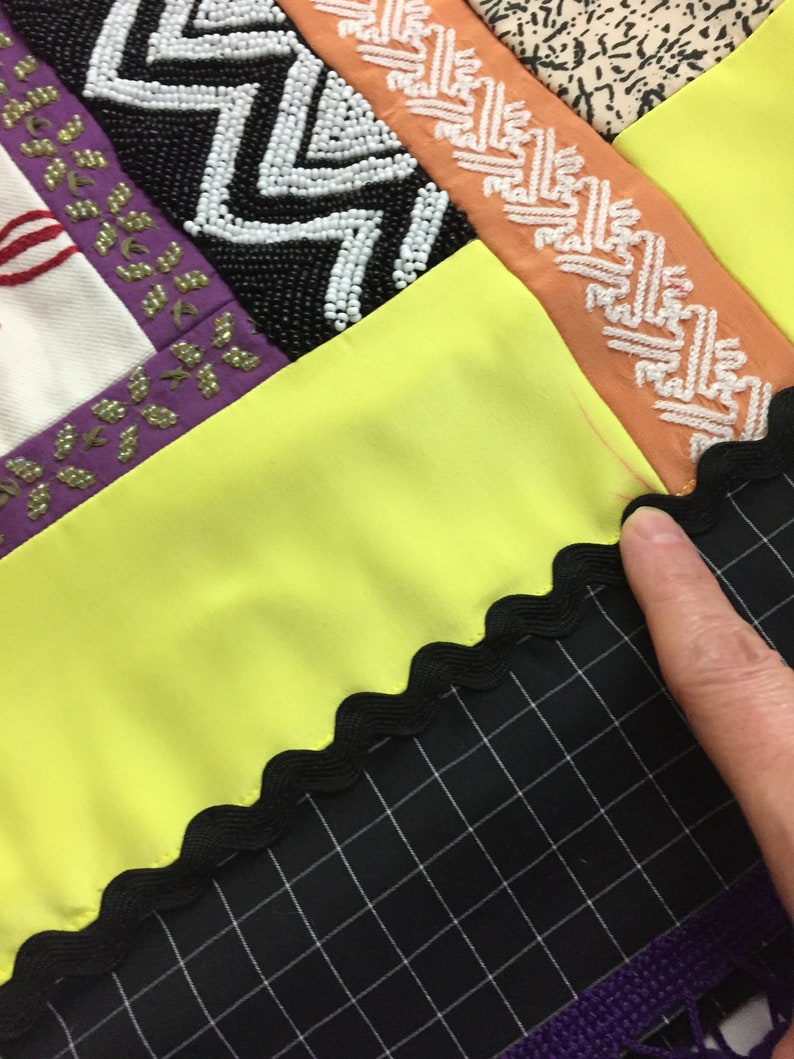 May include: Close-up of a patchwork textile with various textures and colors. Features include black and white beaded chevron patterns, orange and white patterned ribbon, and a bright yellow fabric. A black grid pattern fabric is visible.