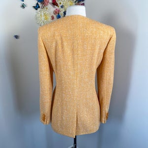 May include: A light orange blazer with a textured fabric and a tailored fit. The blazer has a classic design with a notched collar and buttoned cuffs. The back of the blazer is visible, showcasing the tailored seams and a vent at the bottom.
