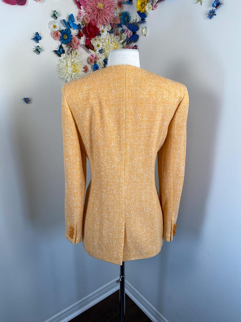 May include: A light orange blazer with a textured fabric and a tailored fit. The blazer has a classic design with a notched collar and buttoned cuffs. The back of the blazer is visible, showcasing the tailored seams and a vent at the bottom.