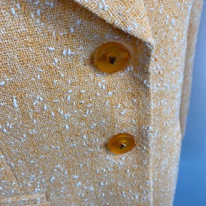 May include: Close-up of a yellow blazer with a textured weave and white flecks. The blazer features two large, round, amber-colored buttons. The fabric has a subtle sheen, suggesting a blend of materials.