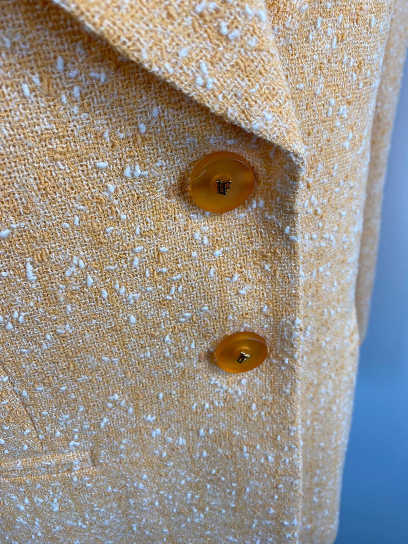 May include: Close-up of a yellow blazer with a textured weave and white flecks. The blazer features two large, round, amber-colored buttons. The fabric has a subtle sheen, suggesting a blend of materials.