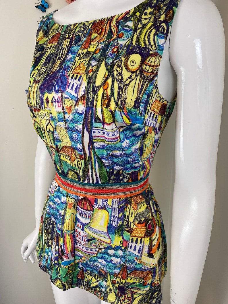 May include: Sleeveless top with a vibrant, artistic print depicting buildings, boats, and figures in blue, yellow, and green hues. The top is accentuated at the waist with a decorative belt. The garment is displayed on a mannequin.