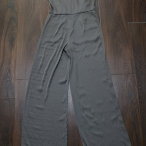 Puede incluir: Mono sin mangas de color verde oliva con cuello alto y perneras anchas. La prenda est&aacute; hecha de una tela suave y cuelga de una percha. Una etiqueta de precio est&aacute; adjunta.