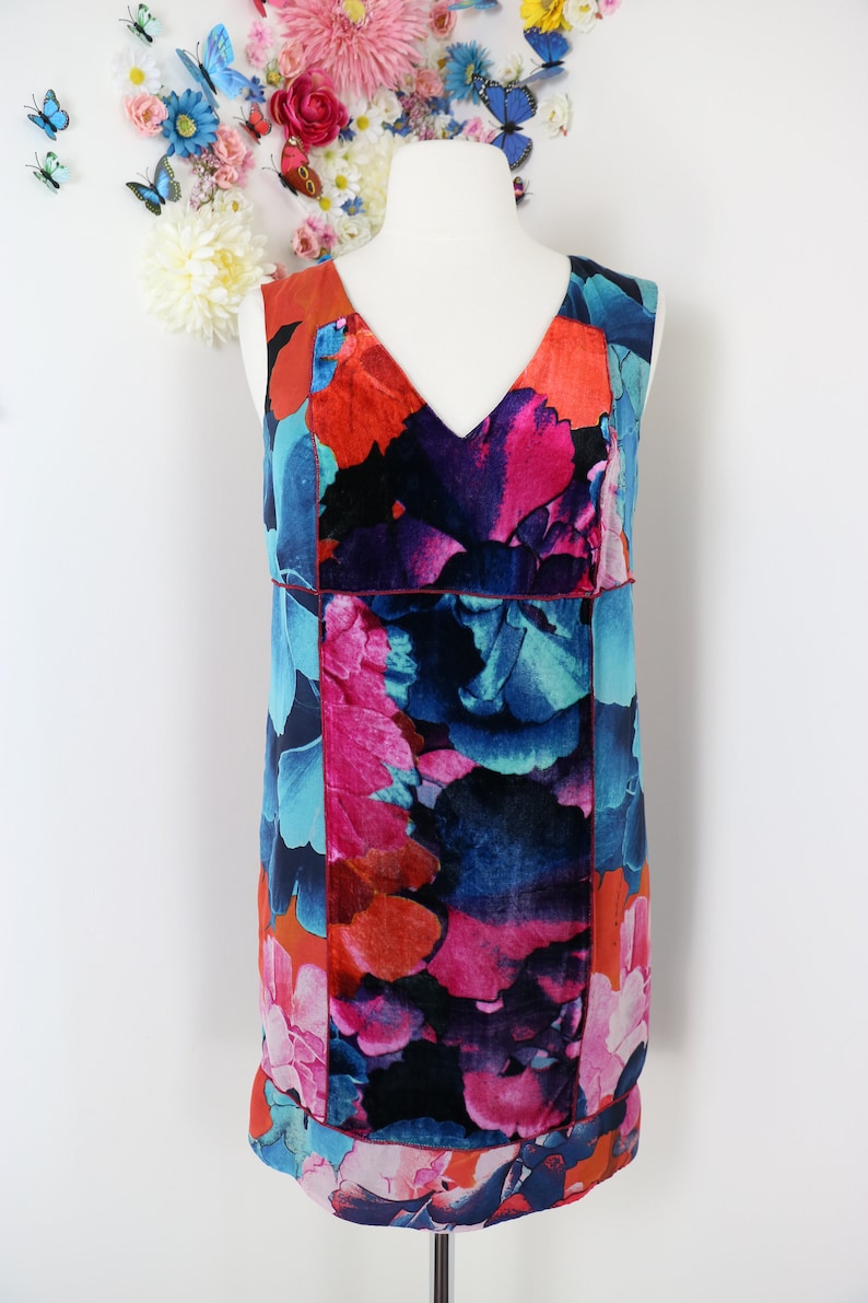 Pode incluir: Vestido sem mangas com decote em V, apresentando um vibrante padr&atilde;o floral em tons de vermelho, rosa, azul e preto. O vestido &eacute; exibido em um manequim sobre um fundo branco, mostrando seu design colorido.