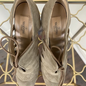 May include: A pair of Valentino Garavani suede high-heeled shoes in a muted olive green. The shoes feature a lace-up design and cut-out detailing. The Valentino Garavani logo is visible on the insole.