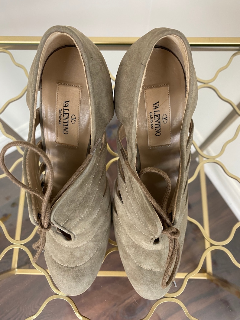 May include: A pair of Valentino Garavani suede high-heeled shoes in a muted olive green. The shoes feature a lace-up design and cut-out detailing. The Valentino Garavani logo is visible on the insole.