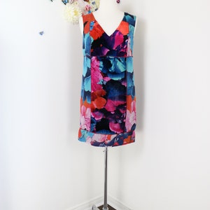 Pode incluir: Vestido sem mangas com decote em V com um padr&atilde;o floral vibrante em tons de vermelho, rosa, azul e preto. O vestido &eacute; exibido em um manequim branco, montado em um suporte cromado. O fundo apresenta uma parede branca com decora&ccedil;&otilde;es florais e borboletas.