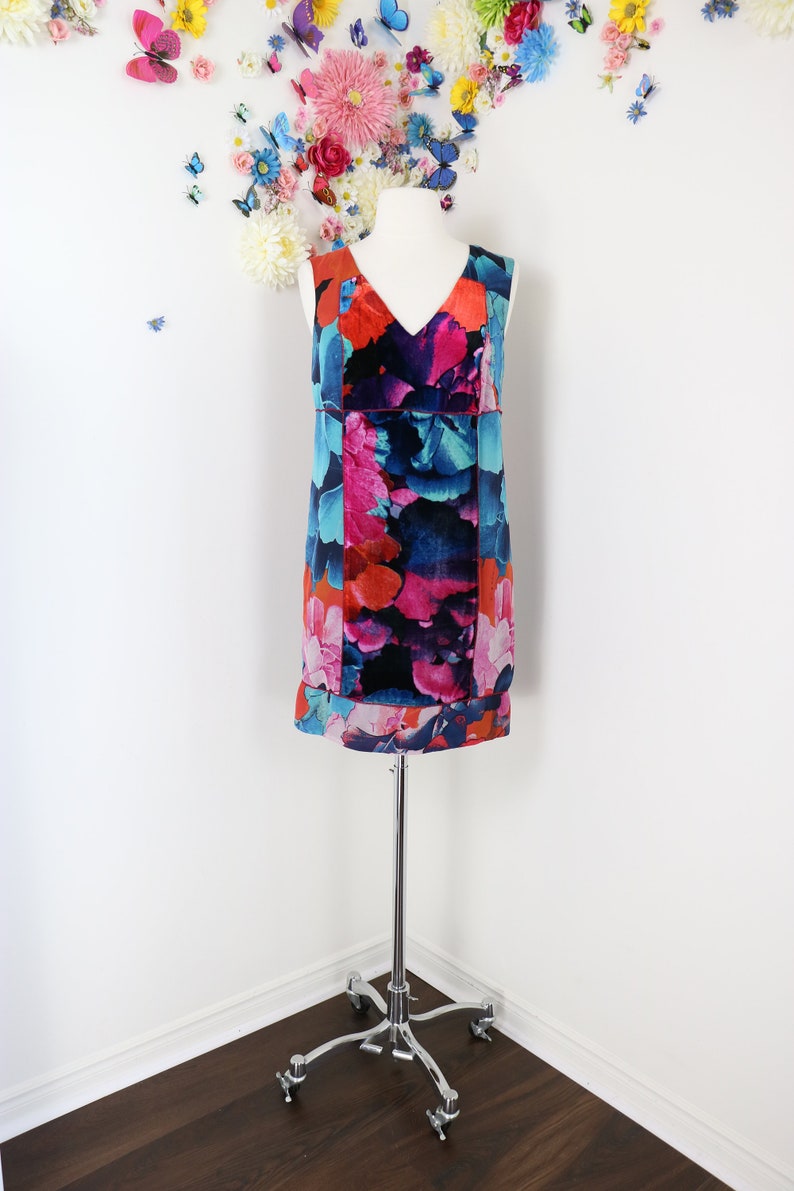 Pode incluir: Vestido sem mangas com decote em V com um padr&atilde;o floral vibrante em tons de vermelho, rosa, azul e preto. O vestido &eacute; exibido em um manequim branco, montado em um suporte cromado. O fundo apresenta uma parede branca com decora&ccedil;&otilde;es florais e borboletas.