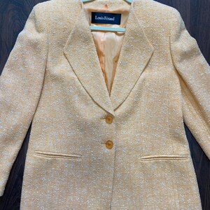 May include: A light orange blazer with a textured fabric, featuring a notched collar and two front pockets. The blazer has two buttons and a label that reads "Louis F&eacute;raud". The jacket is hanging on a light green hanger.