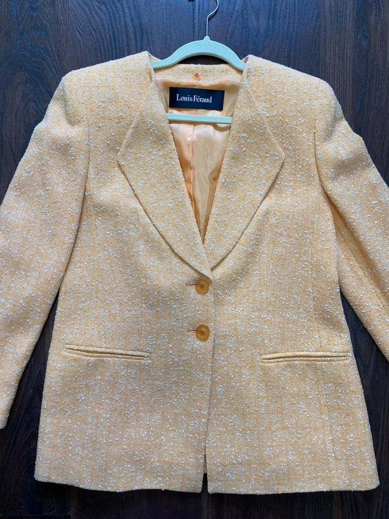 May include: A light orange blazer with a textured fabric, featuring a notched collar and two front pockets. The blazer has two buttons and a label that reads "Louis F&eacute;raud". The jacket is hanging on a light green hanger.