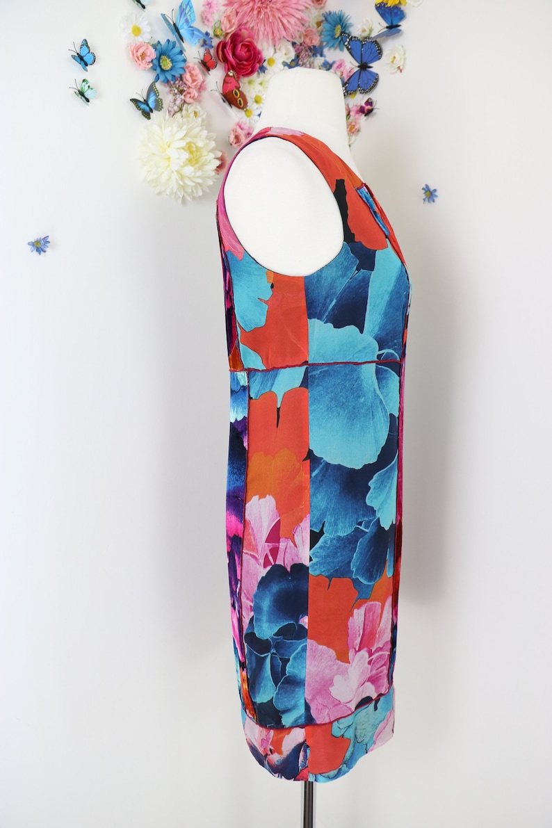 Pode incluir: Vestido sem mangas com um padr&atilde;o floral arrojado em tons de azul, laranja e rosa. O vestido &eacute; apresentado num manequim, destacando o seu design. Tem um decote redondo e um corte reto.