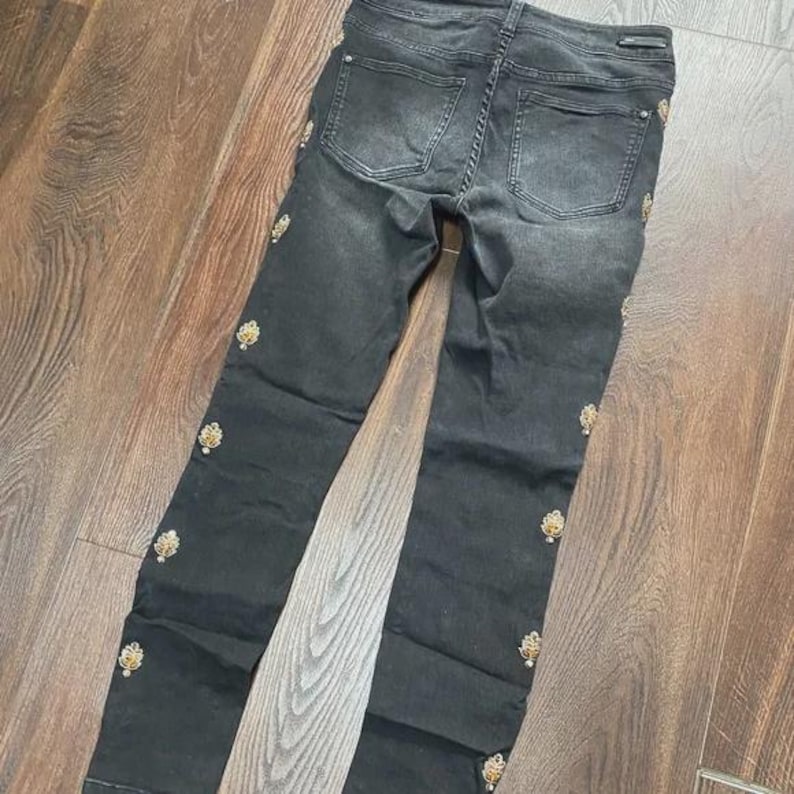 May include: Dark grey denim jeans featuring decorative gold-coloured embellishments along the outer seams. The jeans have a classic five-pocket design and a dark wash. The embellishments add a touch of elegance to the casual style.