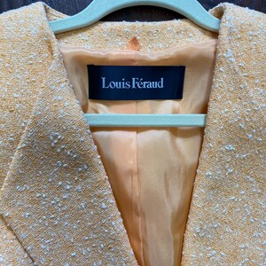 May include: Close-up of a yellow blazer with white flecks, hanging on a green hanger. The blazer has a textured weave and a satin lining. The label inside reads "Louis F&eacute;raud".
