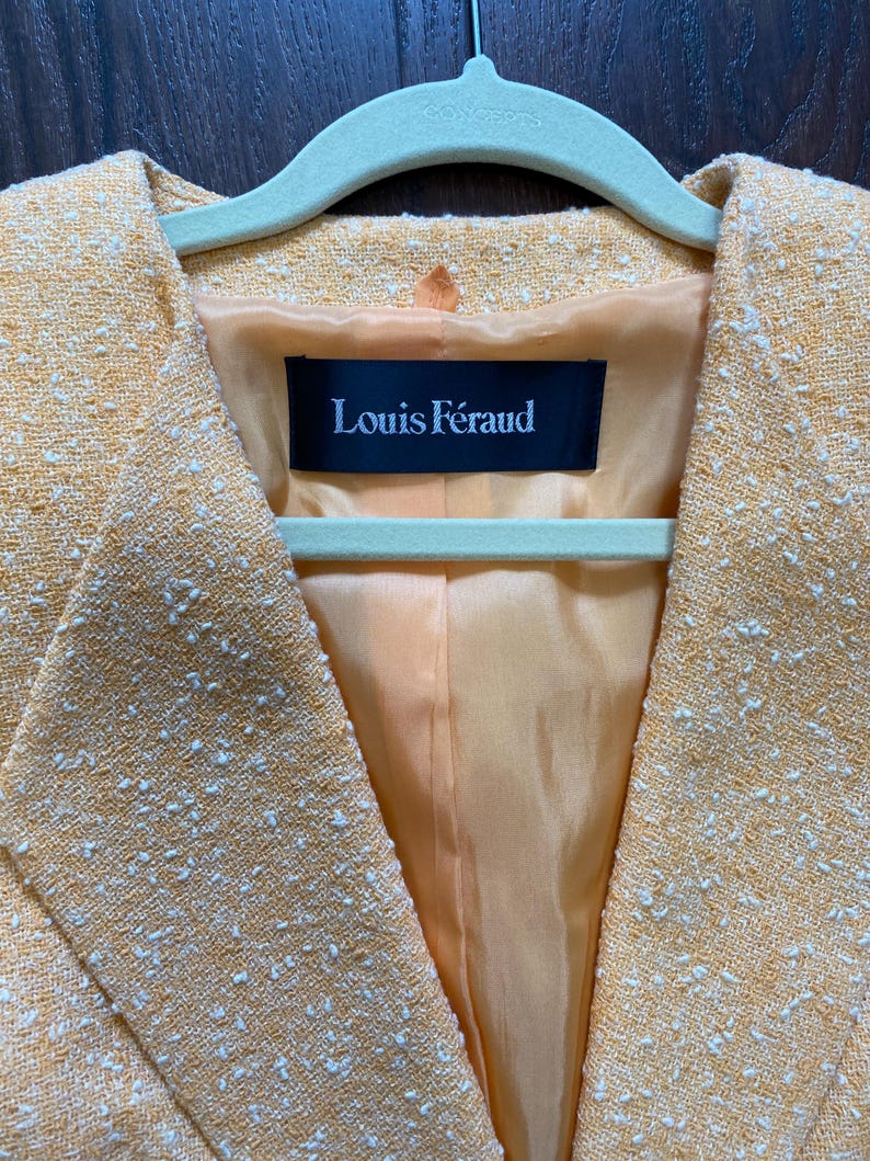 May include: Close-up of a yellow blazer with white flecks, hanging on a green hanger. The blazer has a textured weave and a satin lining. The label inside reads "Louis F&eacute;raud".