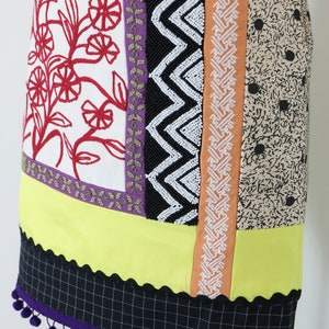 May include: A colorful, patchwork skirt with various patterned panels. The skirt features red floral embroidery, black and white zigzag beading, and a black and white checkered hemline with purple pom-poms. The skirt is displayed on a mannequin.