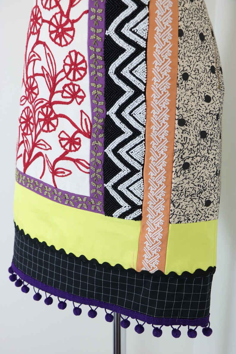 May include: A colorful, patchwork skirt with various patterned panels. The skirt features red floral embroidery, black and white zigzag beading, and a black and white checkered hemline with purple pom-poms. The skirt is displayed on a mannequin.