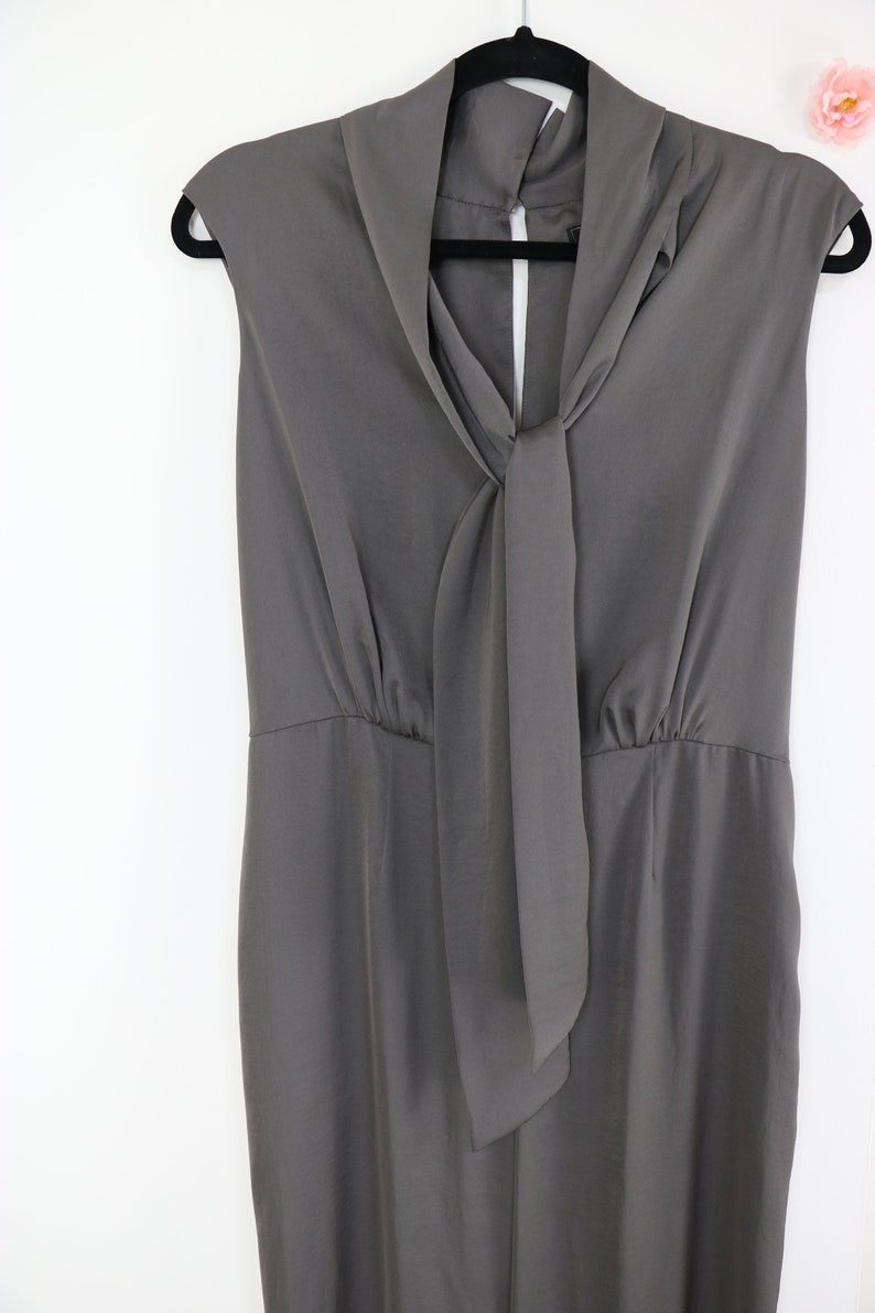 Puede incluir: Un vestido gris sin mangas con cuello drapeado y detalle de lazo. El vestido est&aacute; colgado de una percha negra sobre un fondo blanco. El vestido tiene cintura entallada y falda recta.