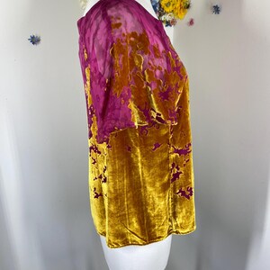 May include: A short-sleeved top featuring a vibrant mustard yellow velvet fabric with a burnout floral pattern that reveals a sheer magenta base layer. The garment has a round neckline and a straight hem, showcasing a textured contrast between the plush velvet sections and the translucent magenta floral design.