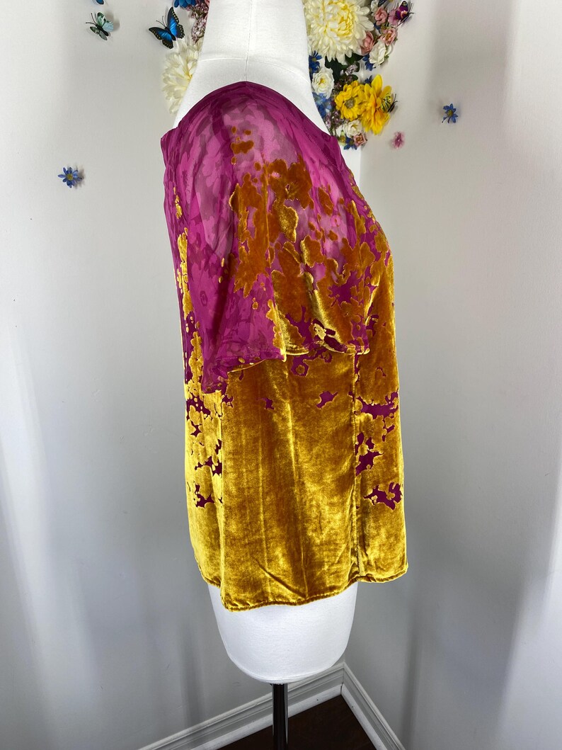 May include: A short-sleeved top featuring a vibrant mustard yellow velvet fabric with a burnout floral pattern that reveals a sheer magenta base layer. The garment has a round neckline and a straight hem, showcasing a textured contrast between the plush velvet sections and the translucent magenta floral design.