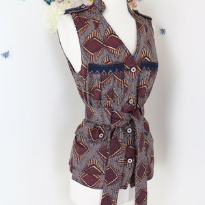 May include: A sleeveless vest with a geometric pattern in shades of brown, burgundy, and gold. The vest features a V-neck, button closures, and a matching tie belt. The vest is displayed on a white mannequin.