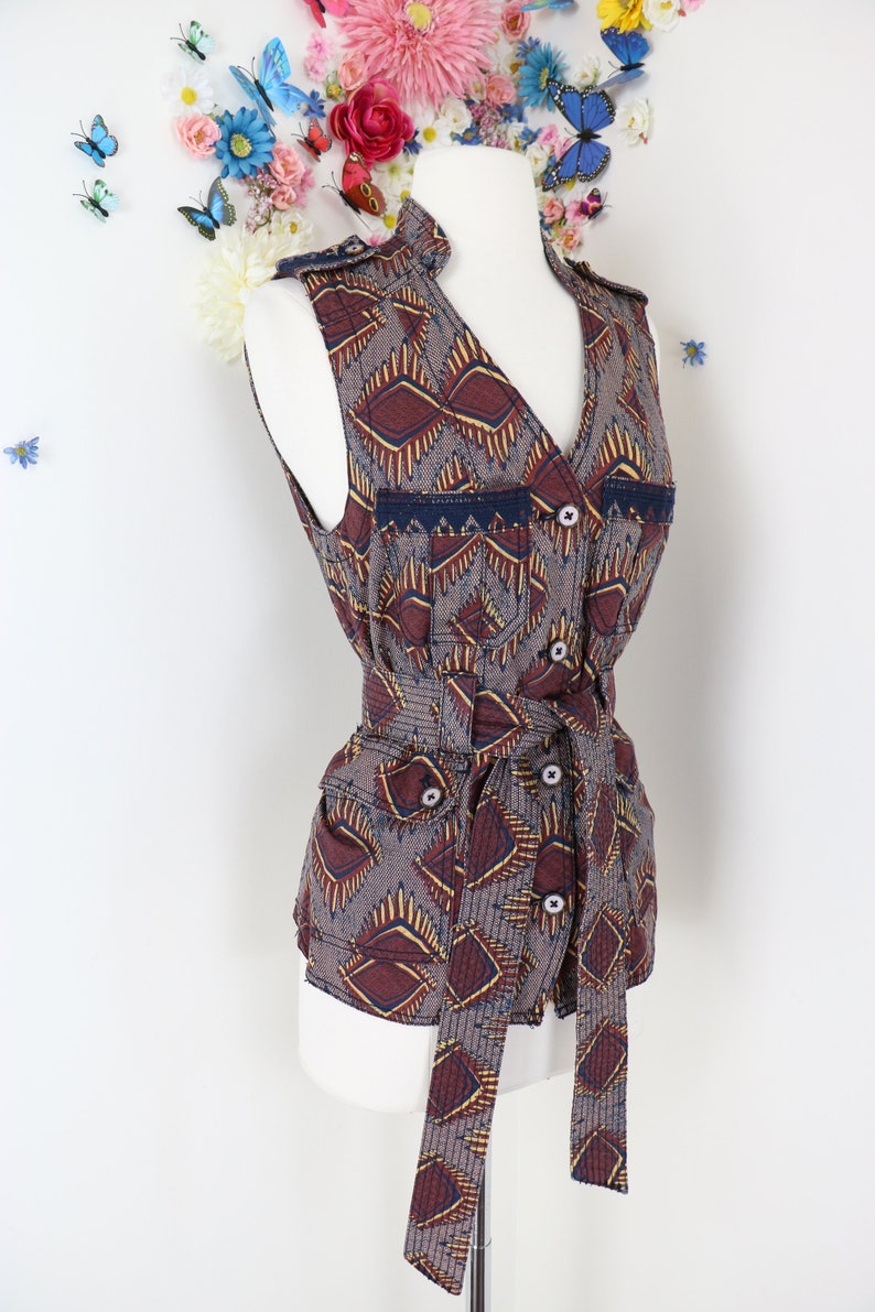 May include: A sleeveless vest with a geometric pattern in shades of brown, burgundy, and gold. The vest features a V-neck, button closures, and a matching tie belt. The vest is displayed on a white mannequin.