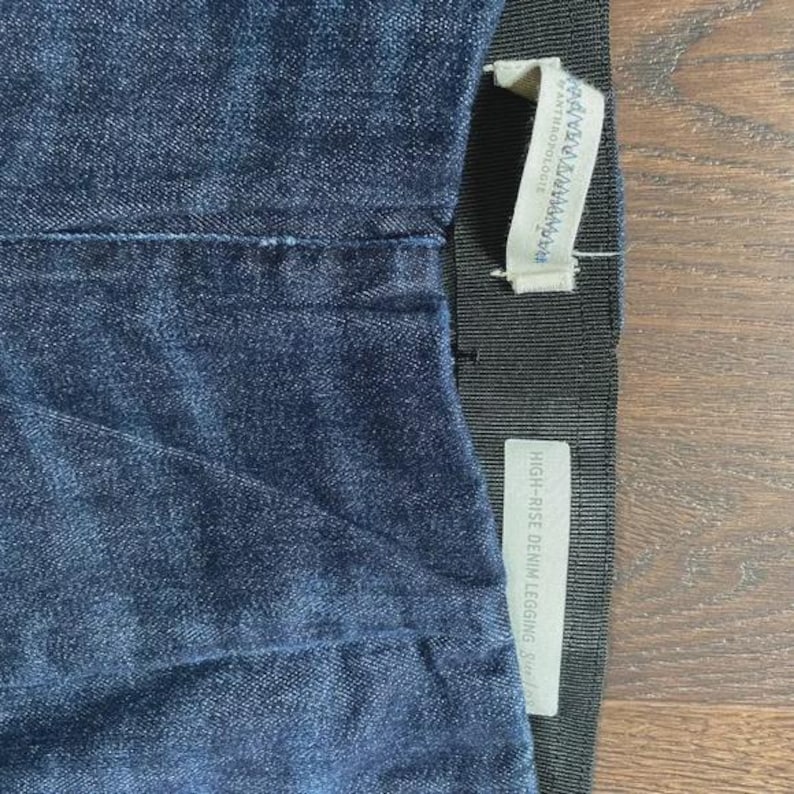 May include: Close-up of dark blue denim leggings. The leggings have a high-rise waist and a black elastic waistband. A white tag with the text "ANTHROPOLOGIE" is attached to the waistband. Another tag reads "HIGH-RISE DENIM LEGGING".