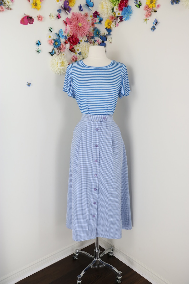 May include: A blue and white striped short-sleeve top paired with a light blue gingham button-down skirt. The skirt has a high waist and is buttoned down the front. The outfit is displayed on a mannequin.