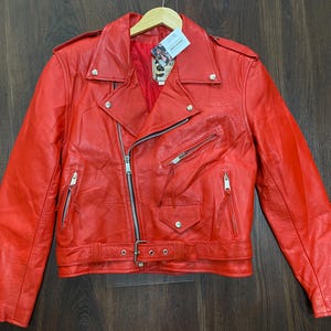May include: A bright red leather motorcycle jacket featuring a wide notched collar, silver-tone metal zippers, and snap button closures. The jacket includes a waist belt with a metal buckle, zippered pockets, and zippered cuffs, all lined in red.
