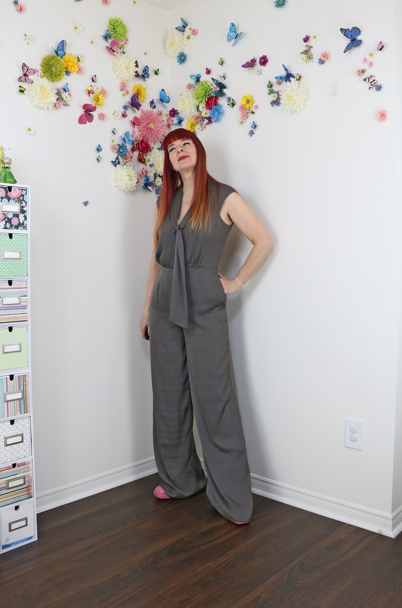 Puede incluir: Mono gris con detalle de cuello anudado y perneras anchas. La prenda sin mangas se lleva con zapatos rosas. Una pared est&aacute; decorada con flores y mariposas de colores.