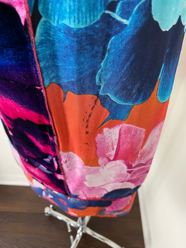 Pode incluir: Um vestido vibrante com estampa floral com grandes flores estilizadas em tons de azul, rosa e laranja. O tecido parece leve, com uma costura vis&iacute;vel no centro. O vestido &eacute; exibido em um manequim prateado.