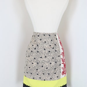 May include: A colorful skirt with a unique design. The skirt features a cream-colored fabric with black polka dots, a yellow scalloped section, and a black and purple checkered hem with purple pom-poms. A white panel with red floral embroidery adds a striking contrast.
