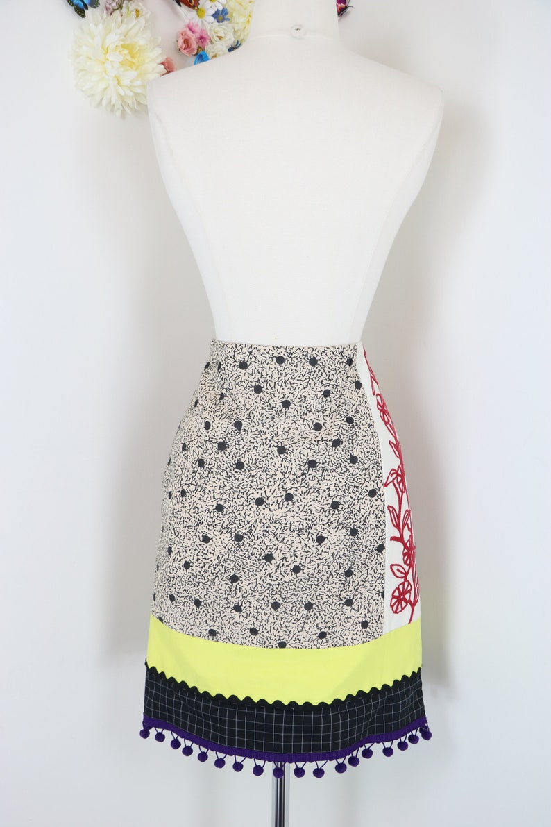 May include: A colorful skirt with a unique design. The skirt features a cream-colored fabric with black polka dots, a yellow scalloped section, and a black and purple checkered hem with purple pom-poms. A white panel with red floral embroidery adds a striking contrast.