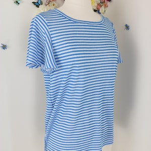 May include: A light blue and white striped t-shirt with short sleeves. The shirt is displayed on a mannequin. The background features a white wall decorated with artificial flowers and butterflies.