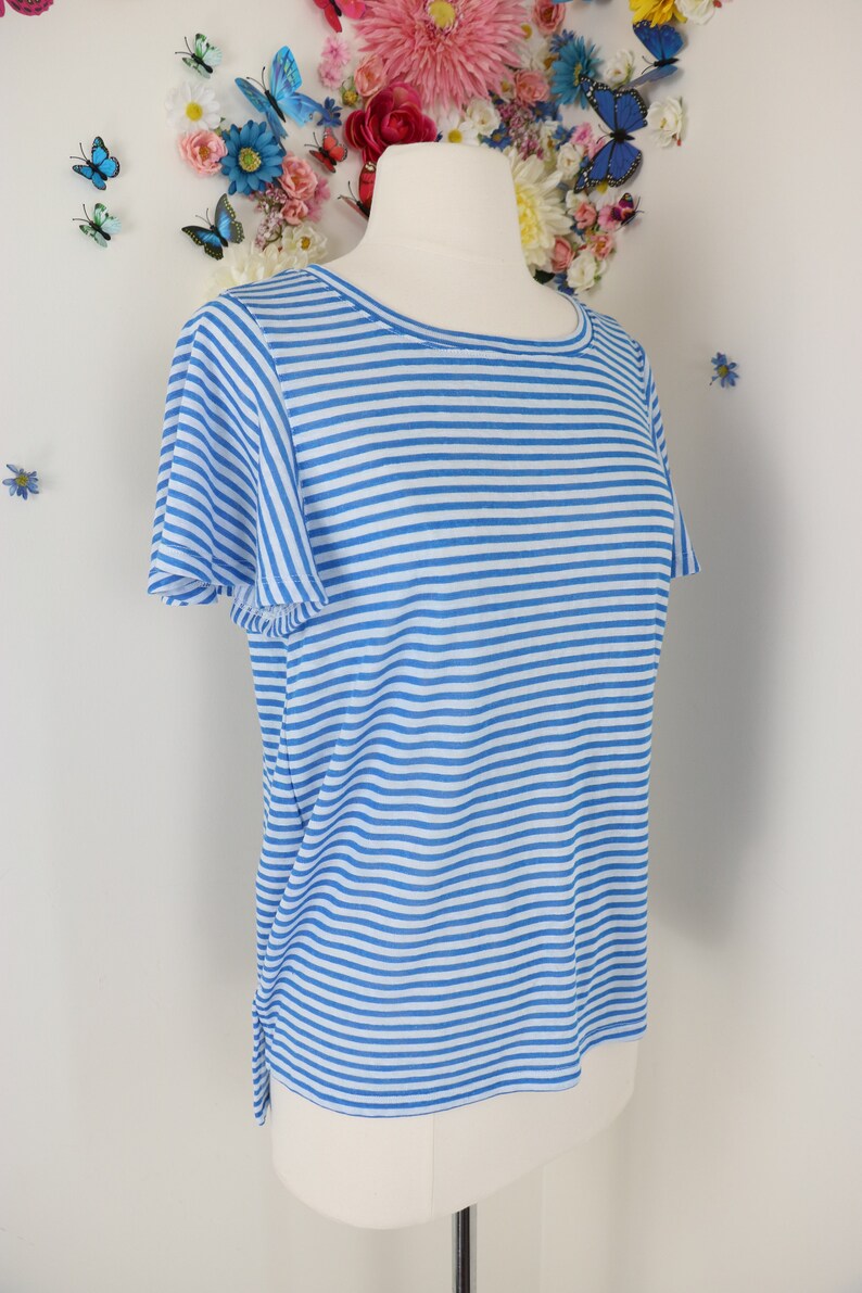 May include: A light blue and white striped t-shirt with short sleeves. The shirt is displayed on a mannequin. The background features a white wall decorated with artificial flowers and butterflies.