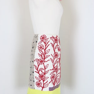 May include: A colorful, patterned skirt with a variety of textures and designs. The skirt features a white background with red floral embroidery, a black and white polka dot panel, and a yellow scalloped hem. The skirt is finished with a black and white checkered panel and purple pom-poms.